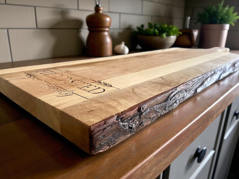May include: A rectangular wooden cutting board with a live edge and the engraved text "FIELDS SHED". The board is light brown with a natural wood grain and a dark brown, textured edge. The board is on a wooden countertop.