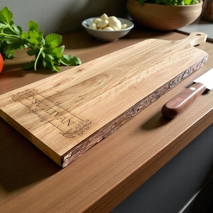 May include: A wooden cutting board with a natural edge and a handle. The board has the engraved text "The O'Sullivan Family" within a decorative frame. A knife with a wooden handle rests on the surface, alongside a tomato and herbs.