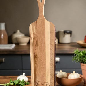 May include: A wooden cutting board with a handle and the text "the FILLDSTED Family". The board is made of light and dark wood tones. A bowl of garlic, a knife, and herbs are in the foreground.