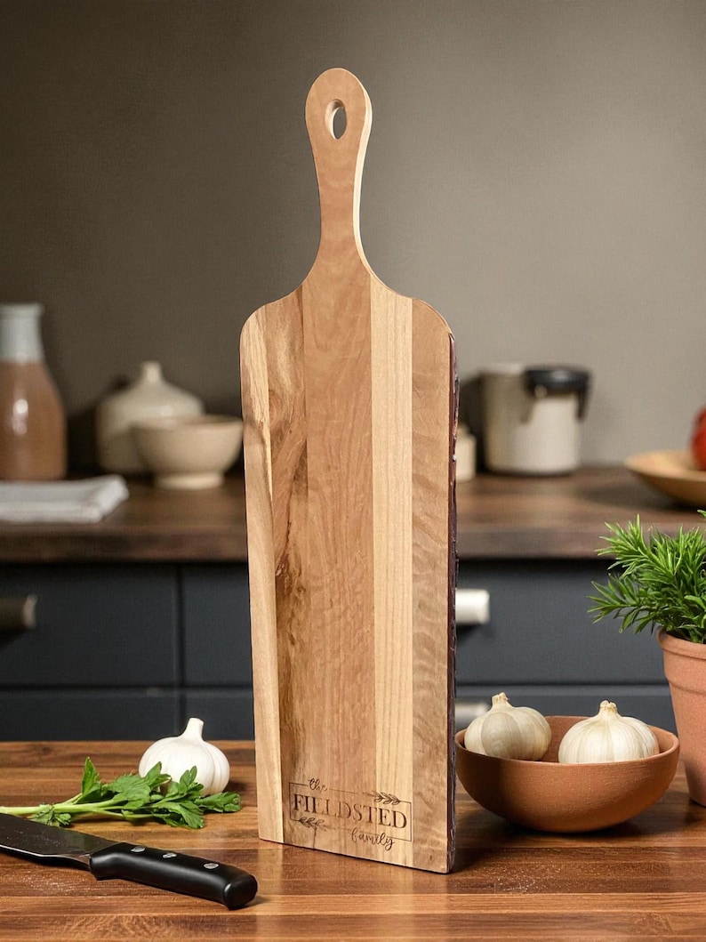 May include: A wooden cutting board with a handle and the text "the FILLDSTED Family". The board is made of light and dark wood tones. A bowl of garlic, a knife, and herbs are in the foreground.