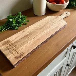 May include: A light-colored wooden cutting board with a handle and live edge. The board has the engraved text "O'Sullivan family" within a decorative frame. Fresh parsley and a bowl of tomatoes are in the background.