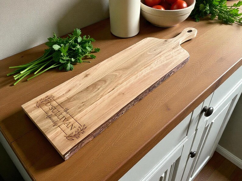May include: A light-colored wooden cutting board with a handle and live edge. The board has the engraved text "O'Sullivan family" within a decorative frame. Fresh parsley and a bowl of tomatoes are in the background.