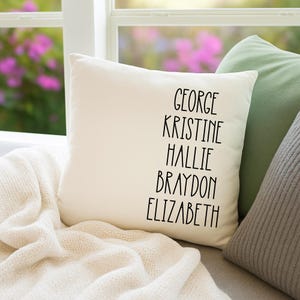 May include: A cream-coloured square cushion with the names George, Kristine, Hallie, Braydon, and Elizabeth printed in black. The cushion rests on a cream-coloured blanket, with green and grey cushions in the background.