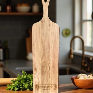 May include: A light-colored wooden cutting board with a handle and a hole for hanging. The board has the text "the O'Sullivan family" engraved on it. The board is rectangular with a natural edge.