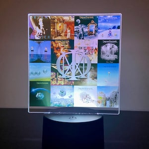 May include: A clear acrylic lamp base with a silver Dream Theater logo in the center. The base features a collage of album covers from the band's discography, including "Scenes from a Memory", "Train of Thought", and "Six Degrees of Inner Turbulence".