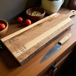 May include: A wooden cutting board with a handle, featuring a natural edge and a light and dark wood grain pattern. The board has the text "the FIELDSTED family" engraved on it. A knife and bowls of food and tomatoes are nearby.