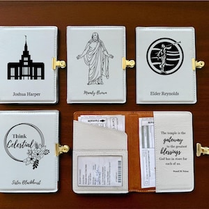 May include: A collection of white leatherette wallets with gold-colored clasps. Each wallet features a unique black graphic and name: a temple, a figure, a circular design, and text. One wallet is open, revealing interior pockets.