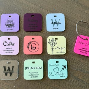 May include: A collection of personalized luggage tags in various colors, including purple, pink, blue, and clear. Each tag features custom text and designs, such as names, initials, and travel-themed graphics. The tags are square with rounded corners and a hole for attachment.