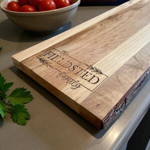 May include: A wooden serving board with a natural edge and a rectangular shape. The board features the engraved text "the FIELDSTED family" within a rectangular frame with leaf accents. The wood grain varies in color.