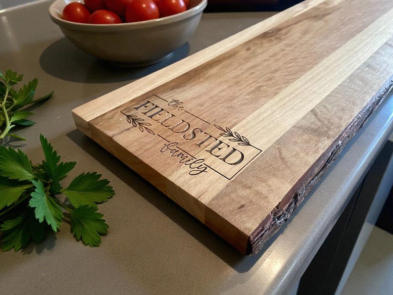 May include: A wooden serving board with a natural edge and a rectangular shape. The board features the engraved text "the FIELDSTED family" within a rectangular frame with leaf accents. The wood grain varies in color.