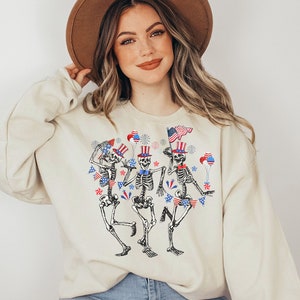 May include: A cream-colored sweatshirt with a graphic of three skeletons wearing red, white, and blue patriotic hats and holding American flags. The skeletons are dancing and surrounded by stars, fireworks, and bunting.