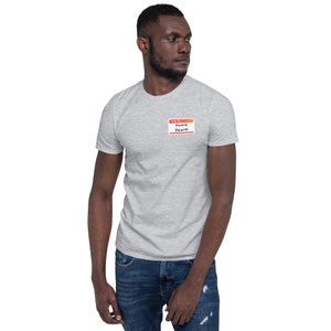 May include: A pale grey t-shirt featuring a name tag that reads "Hello, I'm Howie Dewitt." The name tag has a red border and black text. The t-shirt is short-sleeved.