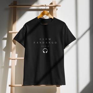 May include: Black t-shirt with the text "CLEM FANDANGO AUDIO" in white. Below the text is a white graphic of headphones. The t-shirt is hanging on a wooden hanger.