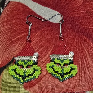 Puede incluir: Un par de pendientes hechos a mano con cuentas que representan una cara de personaje verde con un gorro de Pap&aacute; Noel rojo. Los pendientes tienen ganchos plateados y est&aacute;n colocados sobre un fondo floral rojo. La cara del personaje est&aacute; detallada con cuentas negras, blancas y rojas.