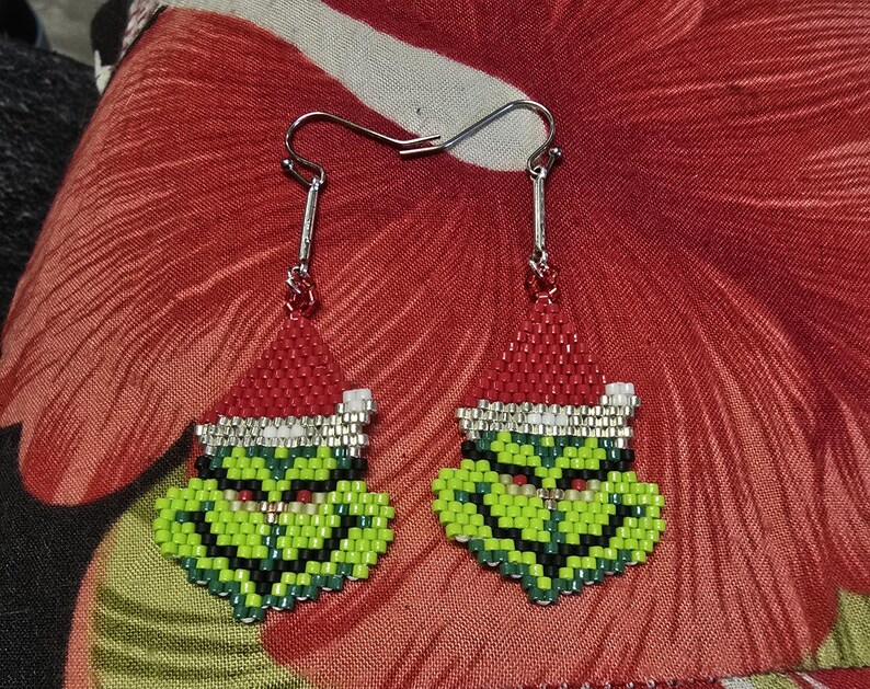 Puede incluir: Un par de pendientes hechos a mano con cuentas que representan una cara de personaje verde con un gorro de Pap&aacute; Noel rojo. Los pendientes tienen ganchos plateados y est&aacute;n colocados sobre un fondo floral rojo. La cara del personaje est&aacute; detallada con cuentas negras, blancas y rojas.