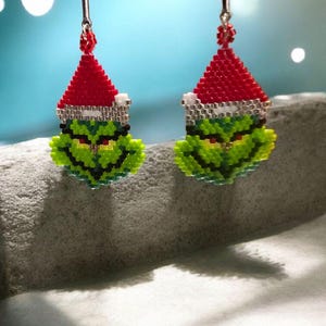 Puede incluir: Un par de pendientes hechos a mano con cuentas que representan la cara del Grinch con un gorro de Pap&aacute; Noel rojo con ribete blanco. Los pendientes son verdes, rojos, blancos y negros, con alambres plateados. Un accesorio festivo navide&ntilde;o.