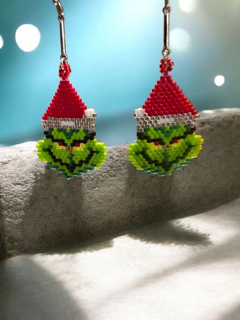 Puede incluir: Un par de pendientes hechos a mano con cuentas que representan la cara del Grinch con un gorro de Pap&aacute; Noel rojo con ribete blanco. Los pendientes son verdes, rojos, blancos y negros, con alambres plateados. Un accesorio festivo navide&ntilde;o.