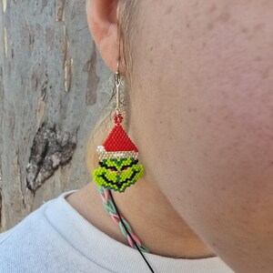 Puede incluir: Un pendiente de cuentas que representa un personaje verde con un gorro de Pap&aacute; Noel rojo. El pendiente tiene un gancho plateado y un cord&oacute;n negro. La cara del personaje es verde con detalles negros, y el gorro es rojo con un borde dorado.