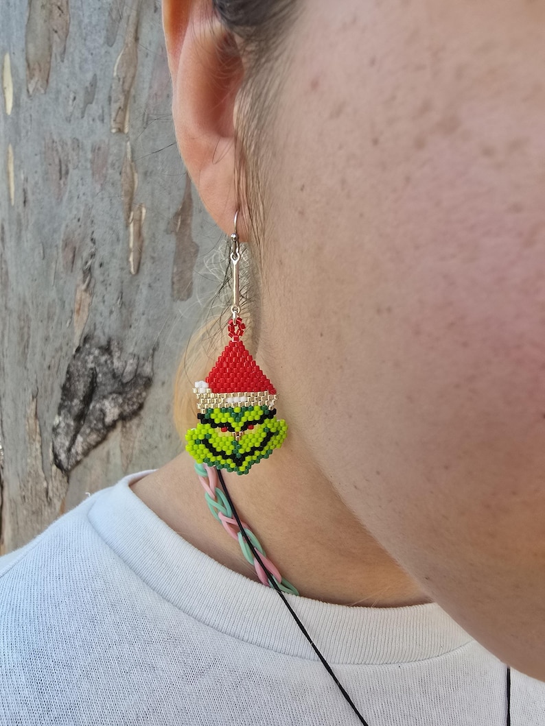 Puede incluir: Un pendiente de cuentas que representa un personaje verde con un gorro de Pap&aacute; Noel rojo. El pendiente tiene un gancho plateado y un cord&oacute;n negro. La cara del personaje es verde con detalles negros, y el gorro es rojo con un borde dorado.
