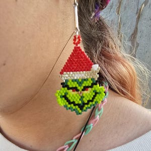 Puede incluir: Un pendiente de cuentas con un personaje verde con un gorro de Pap&aacute; Noel rojo. El pendiente est&aacute; hecho de peque&ntilde;as cuentas de colores y cuelga de un gancho plateado. El personaje tiene una expresi&oacute;n gru&ntilde;ona. Un accesorio festivo.