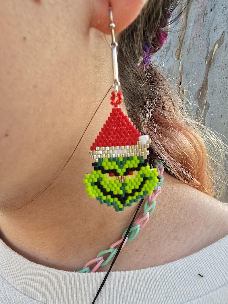 Puede incluir: Un pendiente de cuentas con un personaje verde con un gorro de Pap&aacute; Noel rojo. El pendiente est&aacute; hecho de peque&ntilde;as cuentas de colores y cuelga de un gancho plateado. El personaje tiene una expresi&oacute;n gru&ntilde;ona. Un accesorio festivo.