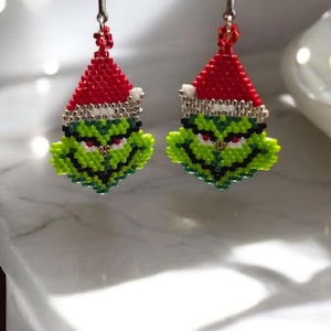 Puede incluir: Un par de pendientes hechos a mano con cuentas que representan al Grinch. Cada pendiente tiene una cara verde con detalles en negro y blanco, un gorro de Pap&aacute; Noel rojo con ribete blanco y ganchos plateados. Perfectos para Navidad.