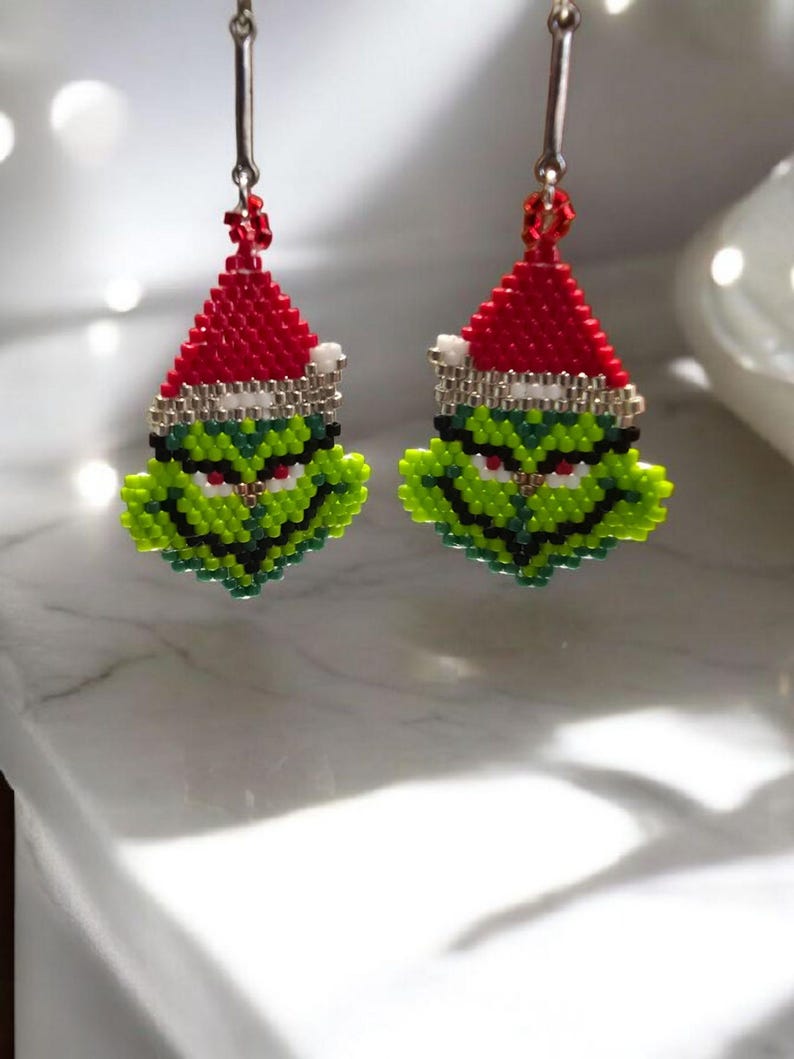 Puede incluir: Un par de pendientes hechos a mano con cuentas que representan al Grinch. Cada pendiente tiene una cara verde con detalles en negro y blanco, un gorro de Pap&aacute; Noel rojo con ribete blanco y ganchos plateados. Perfectos para Navidad.