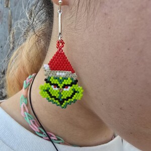 Puede incluir: Un solo pendiente de cuentas con la cara del Grinch con un gorro de Pap&aacute; Noel. El pendiente est&aacute; hecho de cuentas rojas, blancas, verdes, negras y amarillas. Cuelga de un gancho plateado. Un accesorio festivo para las fiestas.