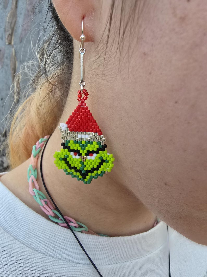 Puede incluir: Un solo pendiente de cuentas con la cara del Grinch con un gorro de Pap&aacute; Noel. El pendiente est&aacute; hecho de cuentas rojas, blancas, verdes, negras y amarillas. Cuelga de un gancho plateado. Un accesorio festivo para las fiestas.