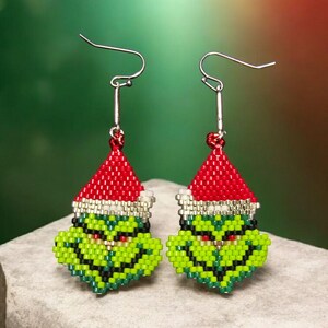 Puede incluir: Un par de pendientes hechos a mano con cuentas que representan una cara de personaje verde con un gorro de Pap&aacute; Noel rojo. Los pendientes tienen ganchos plateados y est&aacute;n sobre un fondo verde y rojo. Un accesorio festivo para las fiestas.
