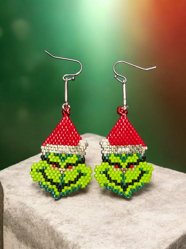 Puede incluir: Un par de pendientes hechos a mano con cuentas que representan una cara de personaje verde con un gorro de Pap&aacute; Noel rojo. Los pendientes tienen ganchos plateados y est&aacute;n sobre un fondo verde y rojo. Un accesorio festivo para las fiestas.