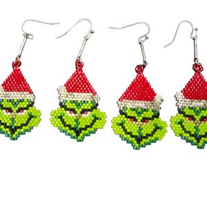 Puede incluir: Cuatro pares de pendientes novedosos con una cara verde con un gorro de Pap&aacute; Noel rojo. Los pendientes est&aacute;n hechos de peque&ntilde;as cuentas y tienen ganchos plateados. La cara tiene una expresi&oacute;n gru&ntilde;ona.