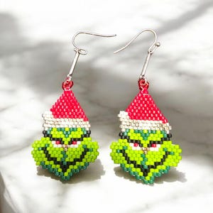 Puede incluir: Un par de pendientes hechos a mano con cuentas que representan una cara verde con un gorro de Pap&aacute; Noel rojo y blanco. Los pendientes tienen ganchos plateados y est&aacute;n dise&ntilde;ados con un tema navide&ntilde;o.
