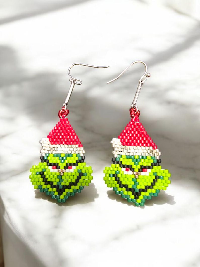 Puede incluir: Un par de pendientes hechos a mano con cuentas que representan una cara verde con un gorro de Pap&aacute; Noel rojo y blanco. Los pendientes tienen ganchos plateados y est&aacute;n dise&ntilde;ados con un tema navide&ntilde;o.