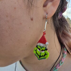 Puede incluir: Un pendiente de cuentas con la cara verde del Grinch que lleva un gorro de Pap&aacute; Noel rojo con ribete blanco. El pendiente est&aacute; hecho de peque&ntilde;as cuentas de colores y cuelga de un gancho plateado.