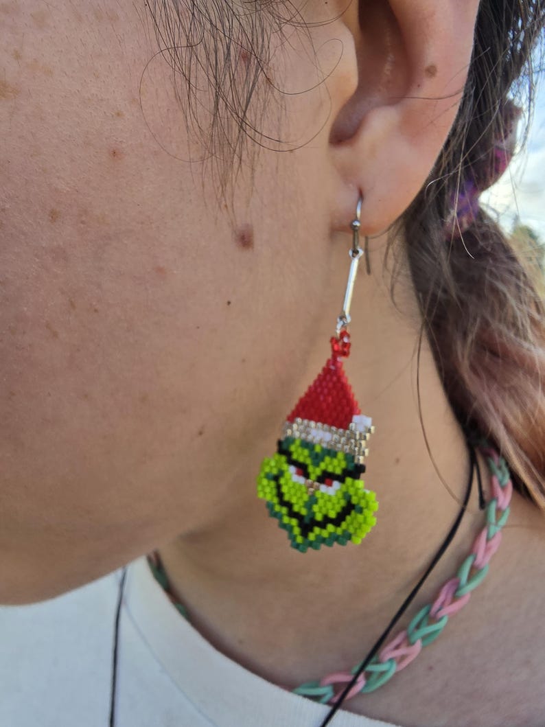 Puede incluir: Un pendiente de cuentas con la cara verde del Grinch que lleva un gorro de Pap&aacute; Noel rojo con ribete blanco. El pendiente est&aacute; hecho de peque&ntilde;as cuentas de colores y cuelga de un gancho plateado.