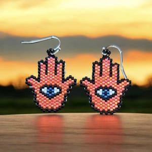 May include: A pair of orange and black beaded earrings shaped like a hamsa hand with a blue evil eye in the center. The earrings are hanging from silver hooks.