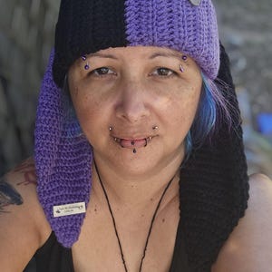 May include: A handmade crochet hat in black and purple, with long ear flaps. The hat has a round emblem on the front and a small tag on the ear flap. The hat is worn by a person with multiple piercings.