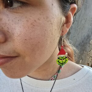 Puede incluir: Primer plano de una oreja con un pendiente tem&aacute;tico navide&ntilde;o. El pendiente presenta una cara verde con un gorro de Pap&aacute; Noel rojo, hecho de peque&ntilde;as cuentas de colores. La persona lleva una camiseta blanca. El pendiente cuelga de un gancho plateado.