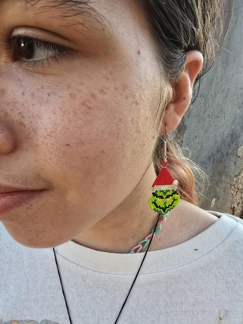 Puede incluir: Primer plano de una oreja con un pendiente tem&aacute;tico navide&ntilde;o. El pendiente presenta una cara verde con un gorro de Pap&aacute; Noel rojo, hecho de peque&ntilde;as cuentas de colores. La persona lleva una camiseta blanca. El pendiente cuelga de un gancho plateado.