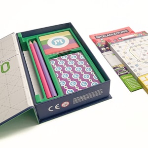 Puede incluir: Una caja azul abierta con un interior verde que contiene un juego. El juego incluye una tarjeta púrpura y azul con un patrón de la letra "M", una tarjeta amarilla, verde y rosa con un símbolo de la letra "M", dos lápices y un conjunto de instrucciones en alemán. La caja tiene una marca CE y la palabra "SPIELANLEITUNG" es visible en las instrucciones.