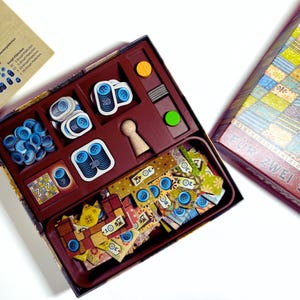Può includere: Un gioco da tavolo con interno marrone, con gettoni blu a forma di bottone, pezzi di gioco in legno e carte da gioco colorate. Il coperchio della scatola recita "FÜR ZWEI". I componenti del gioco sono disposti in una scatola rettangolare.