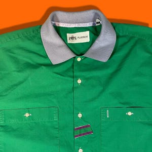 May include: A green button-down shirt with a light blue collar and a Playboy logo on the chest. The shirt has two pockets on the front and a striped patch on the bottom right.