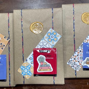 May include: Three brown paper gift bags with decorative paper and wax seals. Each bag has a different tea bag attached with a ribbon. The tea bags are labelled "Blue Butterfly Fleur de pois bleu", "Hibiscus", and "Butterfly".
