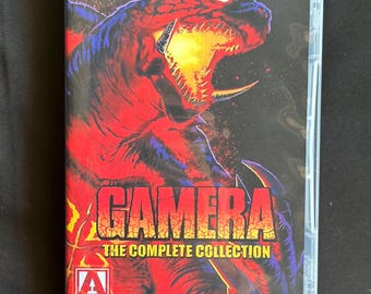 May include: A Gamera The Complete Collection Blu-ray case featuring a large, red and black monster with glowing eyes and an open mouth. The title GAMERA THE COMPLETE COLLECTION is printed in yellow and orange text, with an Arrow Video logo below.