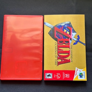 May include: A red plastic video game case next to a rectangular game box for The Legend of Zelda: Ocarina of Time. The gold box features a blue shield and sword graphic, the game title in red text, and the Nintendo 64 logo with an ESRB E rating.