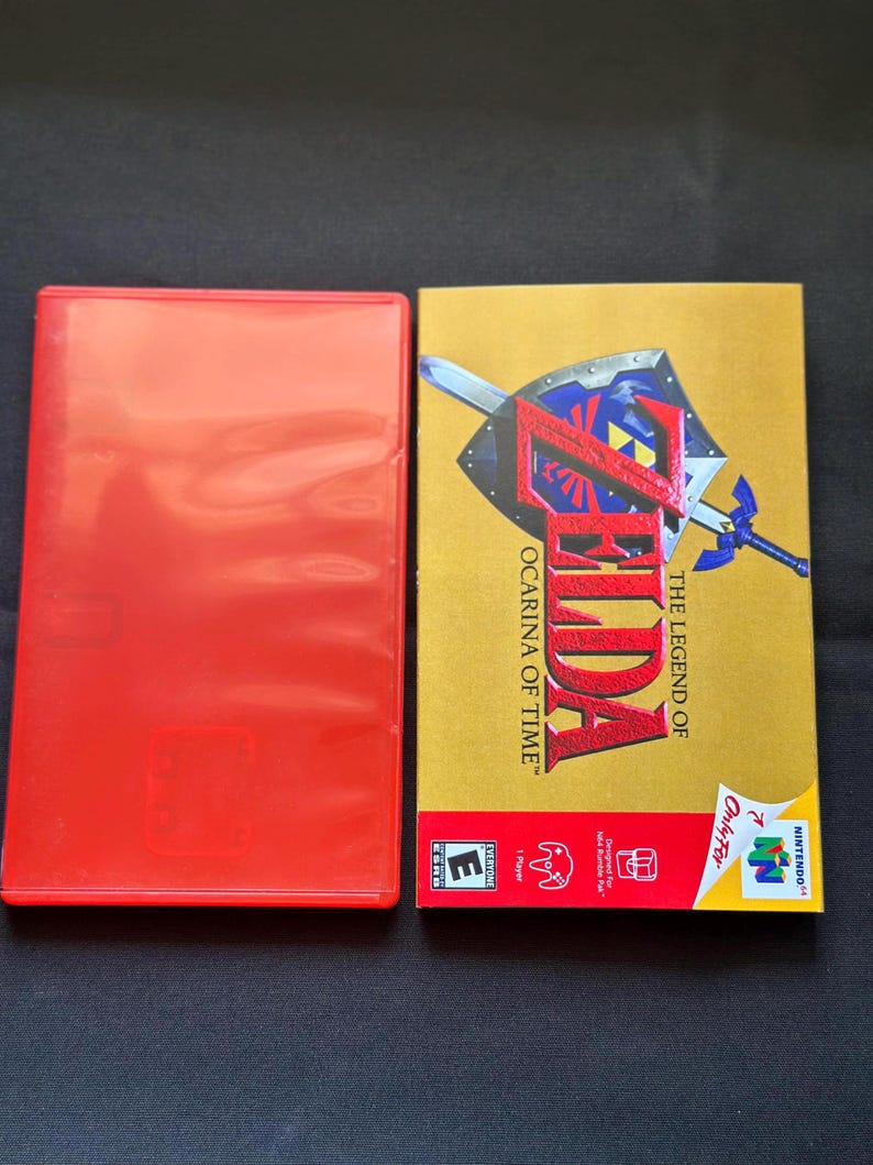 May include: A red plastic video game case next to a rectangular game box for The Legend of Zelda: Ocarina of Time. The gold box features a blue shield and sword graphic, the game title in red text, and the Nintendo 64 logo with an ESRB E rating.