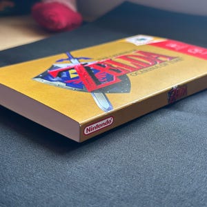 May include: A gold-coloured video game box for The Legend of Zelda: Ocarina of Time. The front cover features a blue and silver shield with a sword and the game title in red text. The Nintendo logo is printed on the side and top corner of the box.