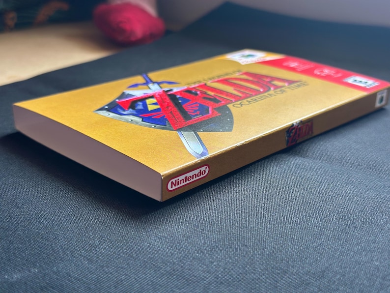 May include: A gold-coloured video game box for The Legend of Zelda: Ocarina of Time. The front cover features a blue and silver shield with a sword and the game title in red text. The Nintendo logo is printed on the side and top corner of the box.
