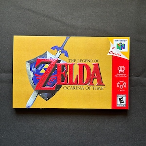 May include: A rectangular gold-colored video game box for The Legend of Zelda: Ocarina of Time for Nintendo 64. The cover features a blue and silver shield with a sword, the red title text, and a red side panel with the Nintendo 64 logo and ESRB rating.